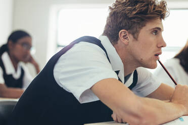 Jugendlicher in Uniform sitzt am Schreibtisch im Klassenzimmer und konzentriert sich. Männlicher Schüler, der im Klassenzimmer aufpasst. - JLPSF23895