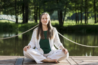 Smiling young woman sitting with cross-legged on pier - LLUF00961