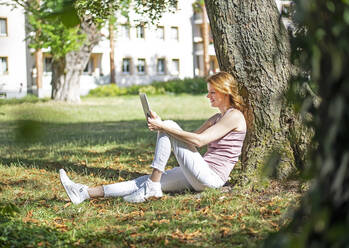 Lächelnde Frau mit Tablet-PC an einem Baumstamm im Park an einem sonnigen Tag - BFRF02419