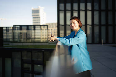 Smiling businesswoman with smart phone standing near railing - MOEF04277