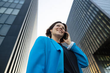 Happy businesswoman talking on mobile phone standing by office building - MOEF04303