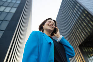 Glückliche Geschäftsfrau, die mit einem Mobiltelefon telefoniert und vor einem Bürogebäude steht - MOEF04303