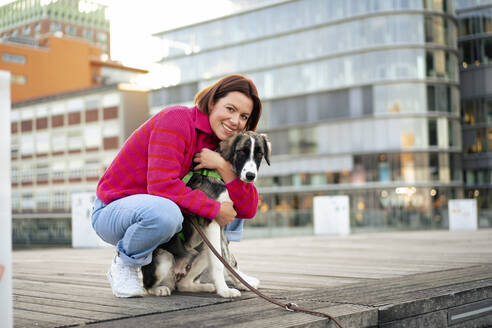 Smiling woman embracing dog crouching on pier - MOEF04339