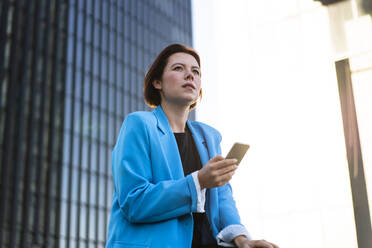 Nachdenkliche Geschäftsfrau mit Smartphone vor einem Bürogebäude stehend - MOEF04356