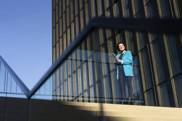 Smiling businesswoman standing near building seen through glass railing - MOEF04367