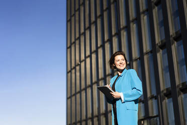 Smiling businesswoman with tablet PC standing near building on sunny day - MOEF04368