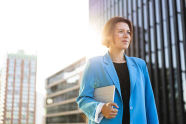 Thoughtful businesswoman wearing blazer holding tablet PC on sunny day - MOEF04375