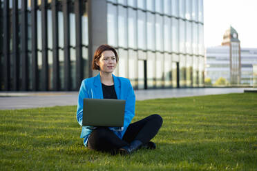 Contemplative businesswoman with laptop sitting cross-legged on grass - MOEF04387