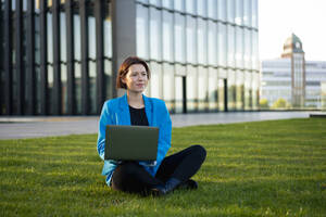 Kontemplative Geschäftsfrau mit Laptop im Schneidersitz im Gras sitzend - MOEF04387