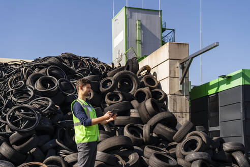 Mann mit Tablet-PC vor Gummireifen im Recyclingzentrum - DIGF19202