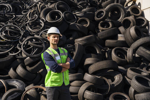 Mann mit Schutzhelm steht mit verschränkten Armen vor Gummireifen in einer Recyclinganlage - DIGF19203