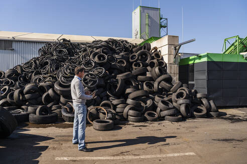 Älterer Geschäftsmann mit Tablet-PC steht neben einem Haufen Gummireifen in einer Recyclinganlage - DIGF19207