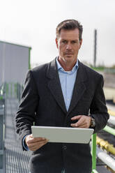 Mature businessman holding tablet PC standing at industrial plant - DIGF19224