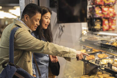 Mature couple shopping pointing at food on display in bakery - JCCMF07775