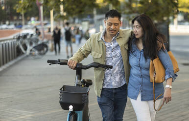 Mature couple walking with electric bicycle on footpath - JCCMF07781
