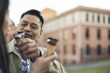 Smiling mature man feeding ice cream cone to woman - JCCMF07785