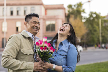 Happy mature couple holding flowers in front of building on Valentine's day - JCCMF07799