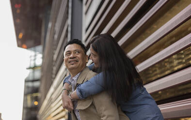 Glückliche Frau umarmt Mann von hinten vor einem Gebäude - JCCMF07809