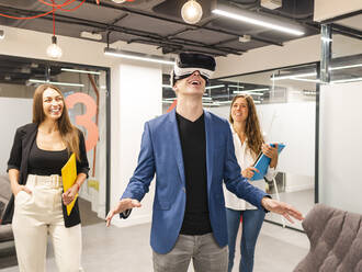 Happy businessman wearing VR glasses with colleagues in background at office - AMRF00134