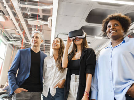 Happy businesswoman wearing VR goggles standing with colleagues in office - AMRF00145