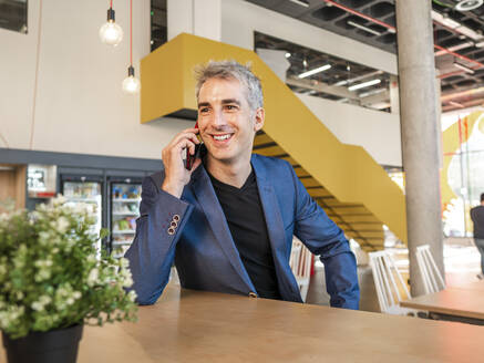 Glücklicher reifer Geschäftsmann, der in einer Cafeteria mit seinem Handy telefoniert - AMRF00155