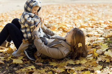 Glückliche Mutter spielt mit ihrem Sohn, der auf Herbstblättern im Park liegt - ONAF00218