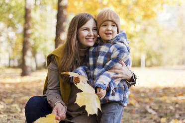 Cute boy with mother holding yellow leaves in park - ONAF00220