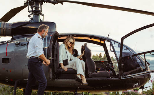 Beautiful young woman getting out of a helicopter with male pilot standing by. Woman travelling by her helicopter. - JLPSF23909