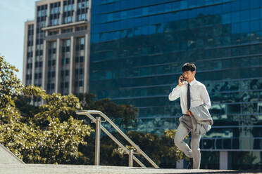 Asian businessman talking on his mobile phone outdoors while walking. Man in businesswear walking up the steps making a phone call against a office building in background. - JLPSF23911