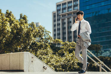 Businessman talking on his cell phone while walking outdoors. Man making a phone call against a office building in background. - JLPSF23912