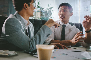 Two business people discussing new project in coffee shop. Young asian business professional explaining some points to his colleague sitting by, at cafe table. - JLPSF23916