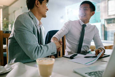 Two successful business men shaking hands with each other after a deal. Asian businesspeople sitting at coffee shop making a hand shake after a successful meeting. - JLPSF23918