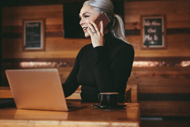 Beautiful senior businesswoman sitting at cafe having telephonic conversation with client. Mature female business professional talking on mobile phone while sitting at coffee shop. - JLPSF23922