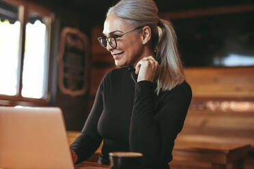 Portrait of smiling senior businesswoman sitting at coffee shop and working on laptop. Beautiful woman at cafe using laptop computer. - JLPSF23928