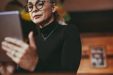 Senior woman wearing eyeglasses reading on digital tablet. Female at cafe using digital tablet. - JLPSF23945