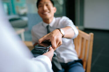 Businessman paying bill through smartphone using NFC technology in restaurant. Closeup of male hand holding his mobile phone over a card reader machine for doing the payment. - JLPSF23950