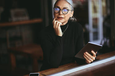 Senior businesswoman sitting at a coffee table with digital tablet. Mature woman relaxing at a restaurant with tablet pc. - JLPSF23959