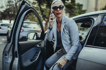 Senior businesswoman getting out of a car talking on phone. Female entrepreneur travelling to office in a luxurious car. - JLPSF23970