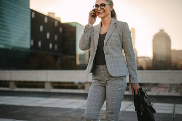 Beautiful senior woman walking outside on road talking on phone. Businesswoman walking outdoors on city street with mobile phone. - JLPSF23982