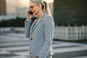 Mature businesswoman on way to home using phone. Senior executive walking on city street talking on cellphone during evening. - JLPSF23984