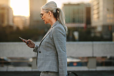 Side view of senior businesswoman reading text message on her smart phone while walking outdoors. Mature woman reading messages on a smartphone while walking on urban background. - JLPSF23985