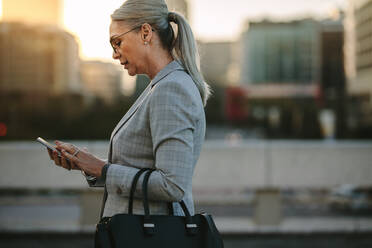 Professional business woman using mobile phone outdoors. Female texting on smart phone while walking outdoor in city. - JLPSF23986