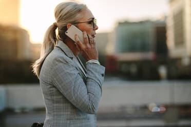 Side view of senior business professional walking outside on road talking on cell phone. Mature businesswoman walking outdoors on street with mobile phone. - JLPSF23987