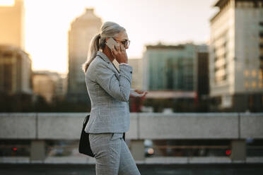 Side view of mature woman in business suit walking back to home talking on phone. Senior businesswoman using cellphone on city street with urban background. - JLPSF23988