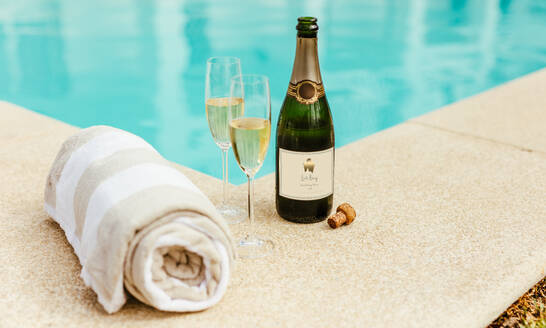 Two glasses of wine with bottle of sparkling wine and soft cotton towel on side of a swimming pool edge. - JLPSF23993