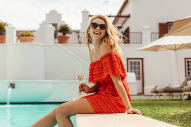 Smiling woman sitting on the pool edge with glass of wine. Beautiful woman relaxing outdoors by swimming pool. - JLPSF23999