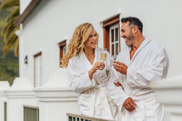 Beautiful couple in bathrobe clinking glasses of wine and smiling. Cheerful couple wearing bathrobes with glasses of champagne in luxury villa. - JLPSF24010