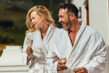 Man and woman in bathrobe with glasses of wine looking away and smiling. Couple wearing bathrobes standing in luxury villa balcony. - JLPSF24013