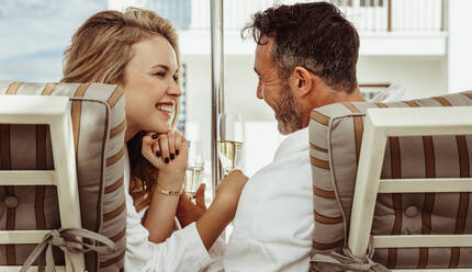 Romantic couple in bathrobes looking at each other with a glass of wine in hand. Smiling man and woman sitting on deck chairs outdoors. - JLPSF24017