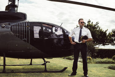 Mature male pilot in uniform standing outside with a private helicopter. Private helicopter parked in the lawn with male pilot waiting. - JLPSF24019